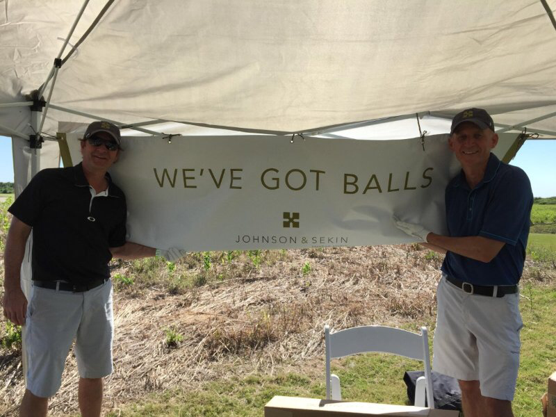 Johnson and Sekin proudly dispaly company booth at golf tournament with sign that reads, 